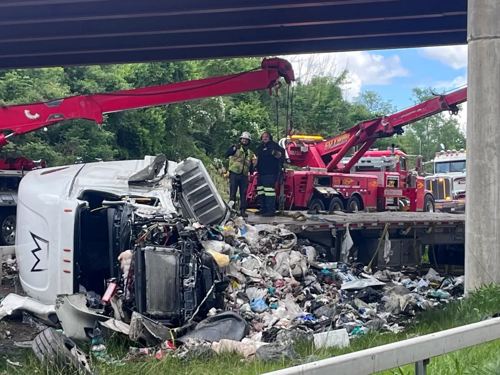 Citywide recovery units at an overturned tractor-trailer with spilled cargo under a highway overpass